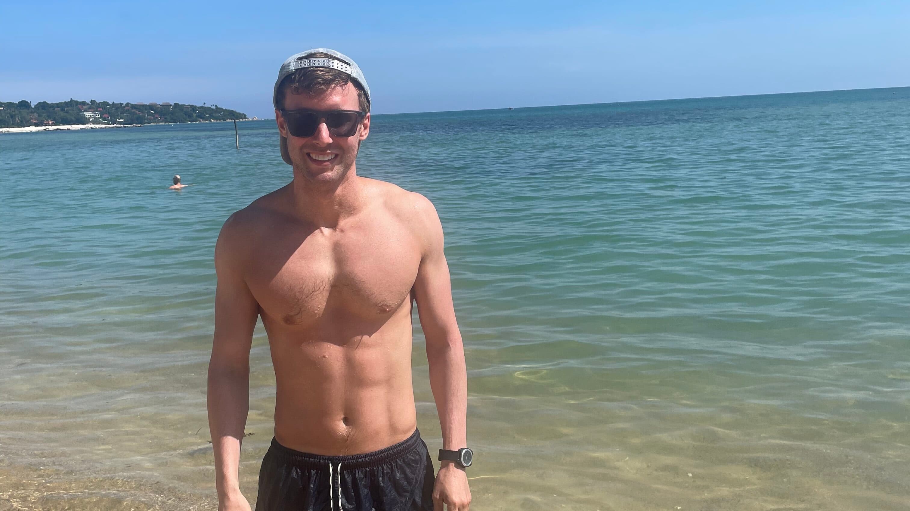 Andrew on beach standing on sand sea in the back with hat and sunglasses on in swimming shorts