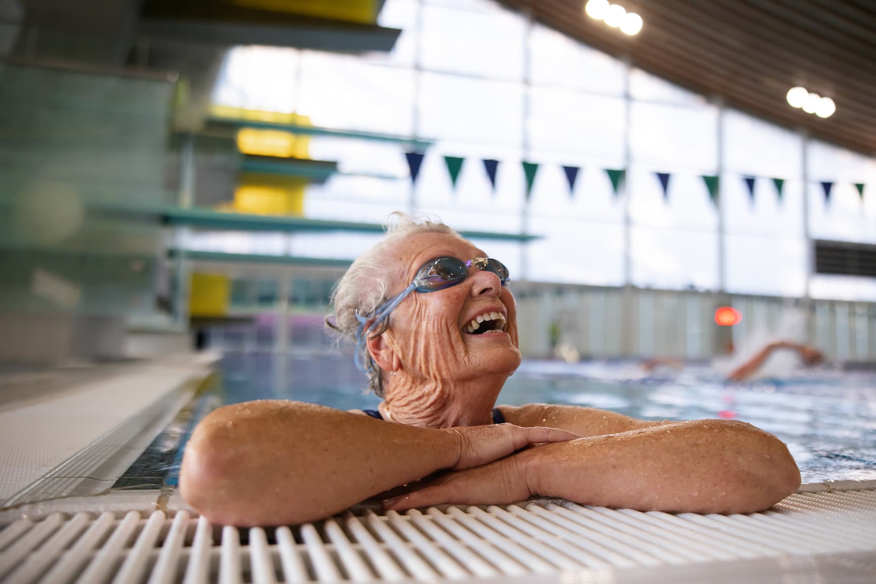 Older woman wearing swim goggles smiling while resting at the edge of an indoor swimming pool, representing active aging and wellness.