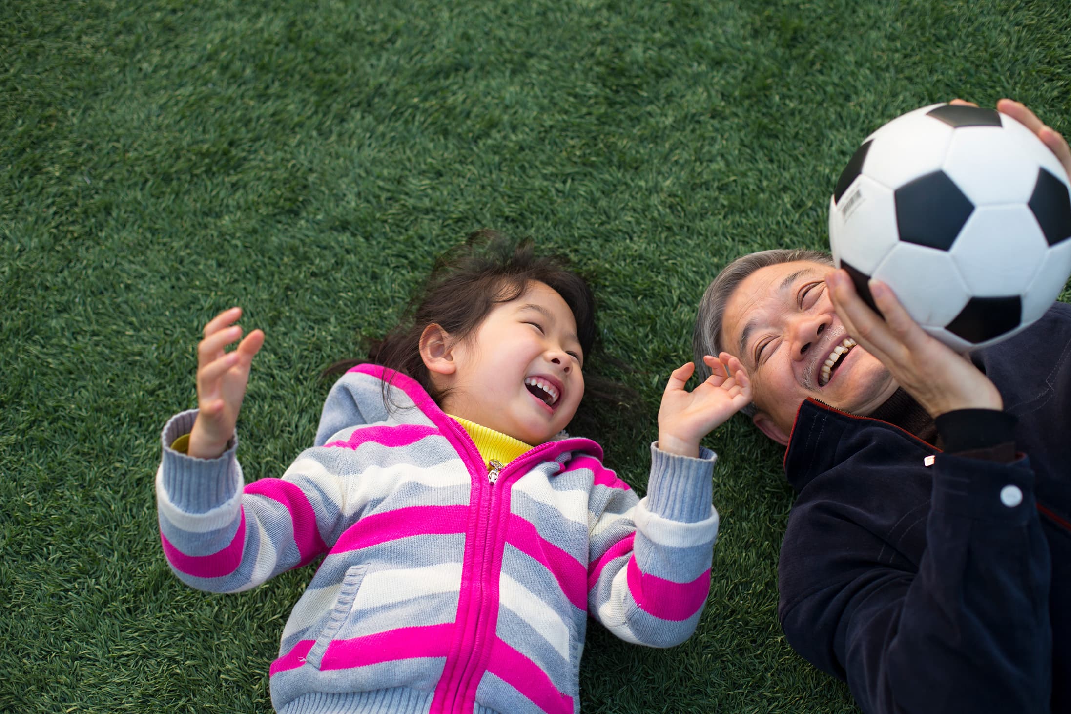 Grandparent and young child lying on green grass laughing while playing with a football, capturing joyful family bonding, outdoor play, and intergenerational connection.
